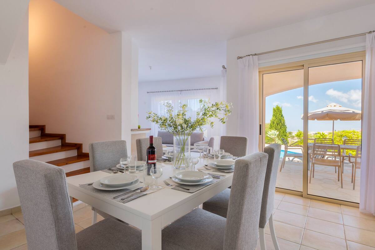 A dining area with a white table set for four, a vase with flowers, and a view of the outdoor patio with a pool and umbrellas.
