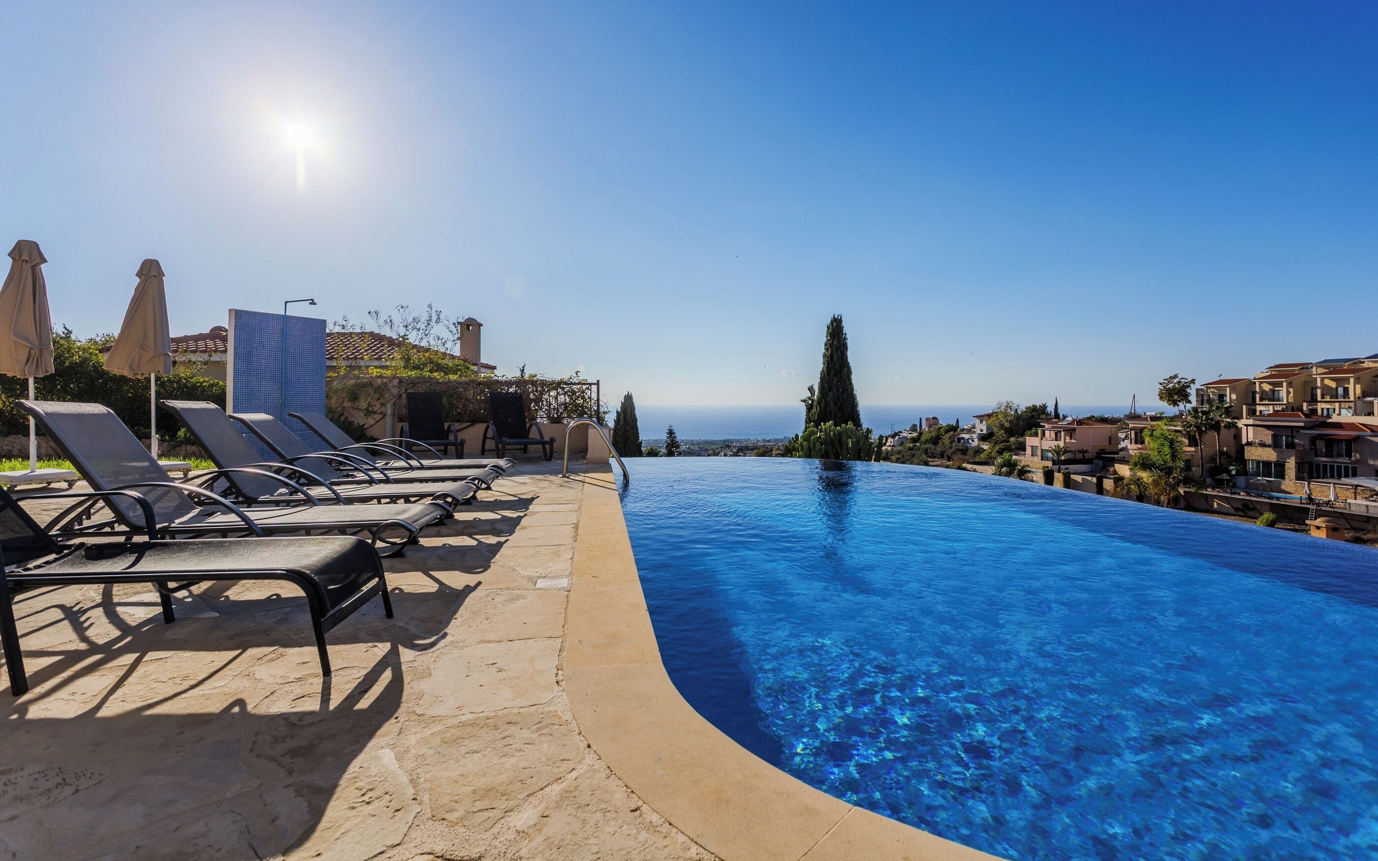 A clear blue infinity pool with lounge chairs and umbrellas, overlooking a coastal town.