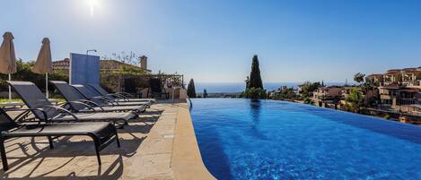 A clear blue infinity pool with lounge chairs and umbrellas, overlooking a coastal town.