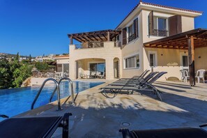 Vista de la piscina con sillas de descanso, cielo despejado y un edificio con paredes de piedra y balcones.