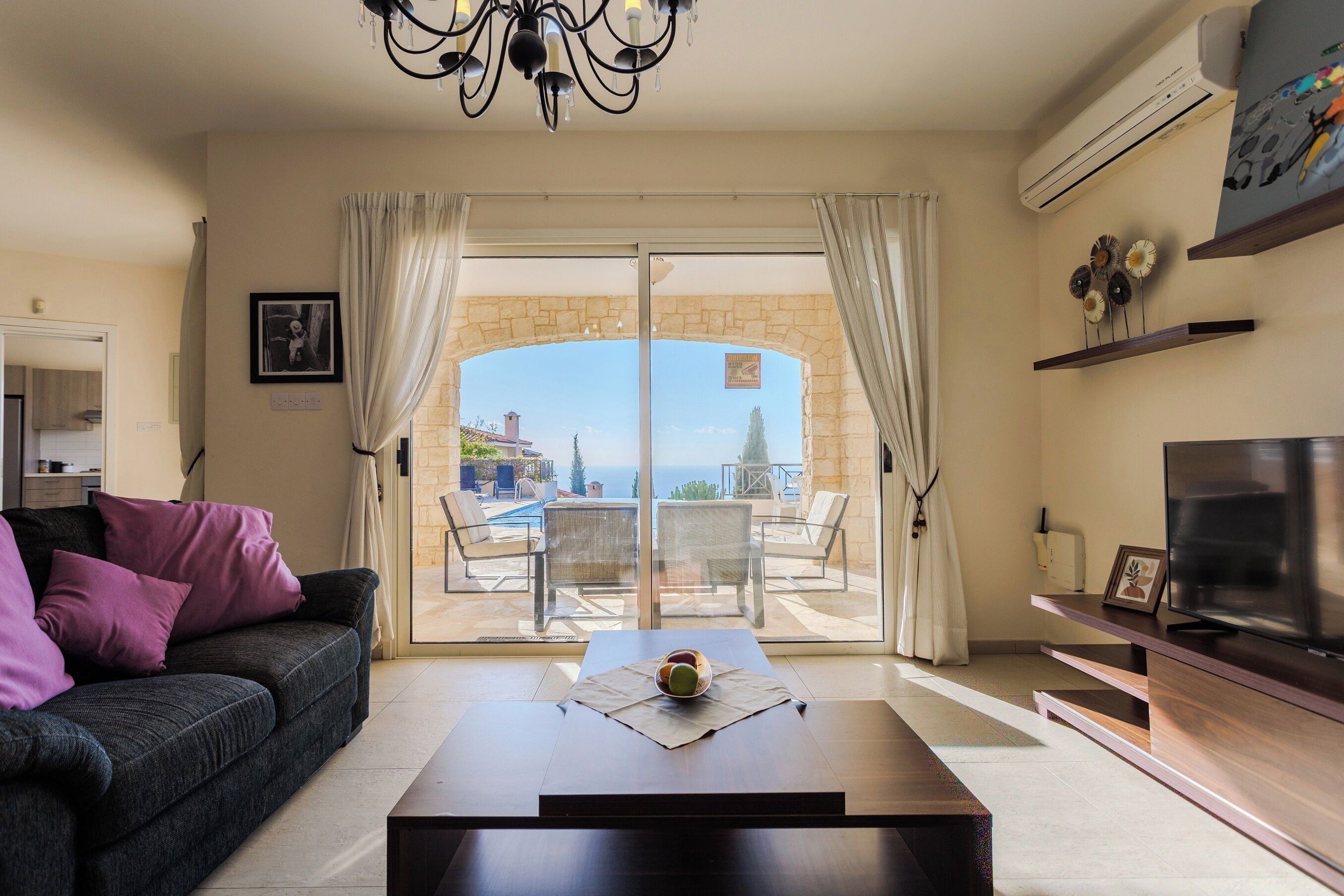 A living room with a sofa, a coffee table, and a TV. There is a view of a pool and a stone wall.