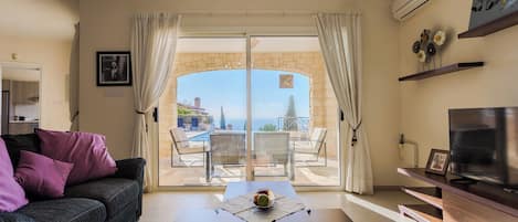 A living room with a sofa, a coffee table, and a TV. There is a view of a pool and a stone wall.