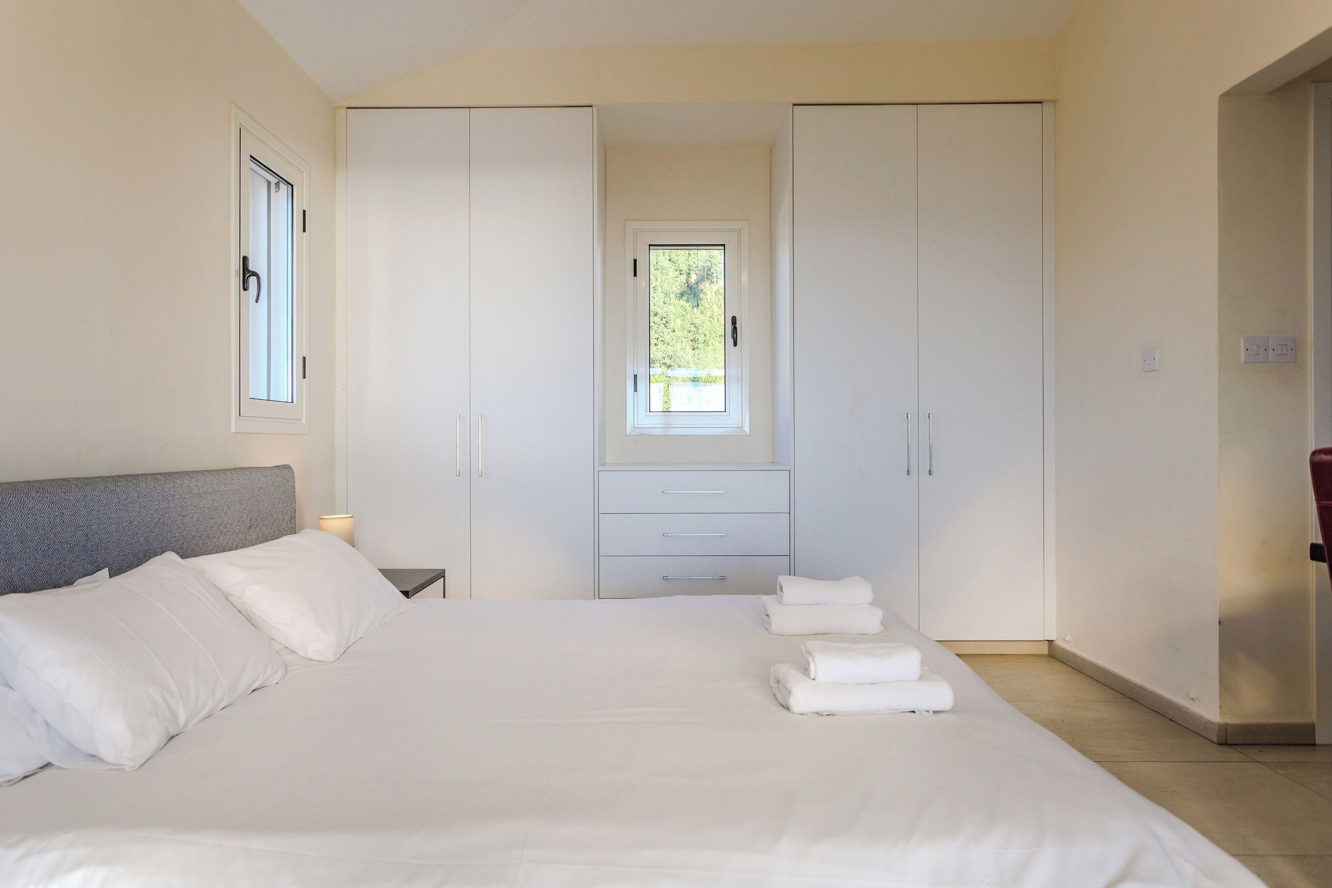 Un dormitorio con una cama grande, un armario, una mesita de noche con lámpara y una ventana con vistas a zonas verdes.