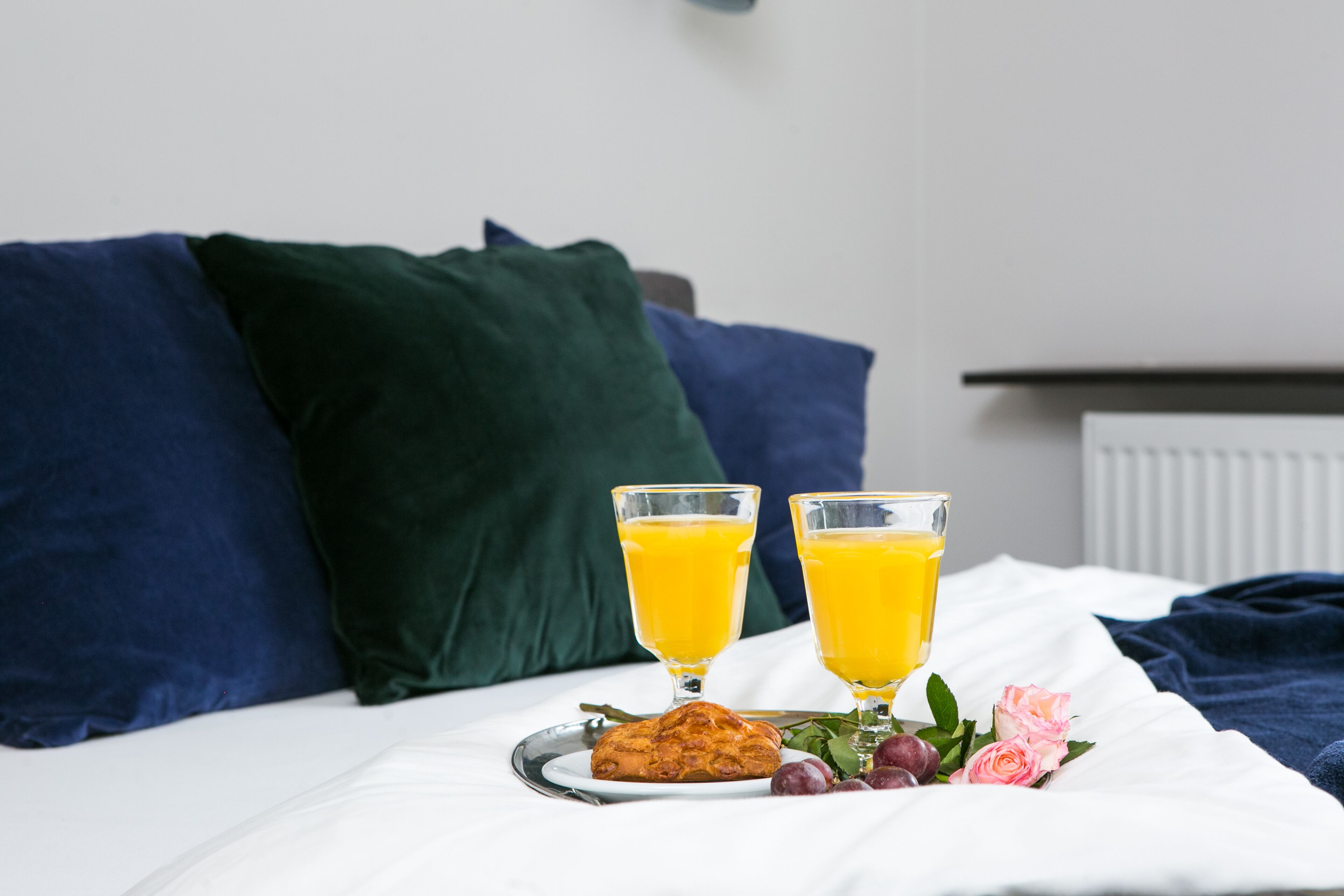 A neatly made bed with two glasses of orange juice, a plate with a muffin and grapes, and a bouquet of pink roses on a white bedspread.