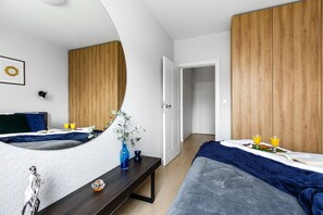 Une chambre moderne avec un grand miroir, une armoire en bois, une table de chevet ornée d’un vase et un lit recouvert d’une couverture bleue.