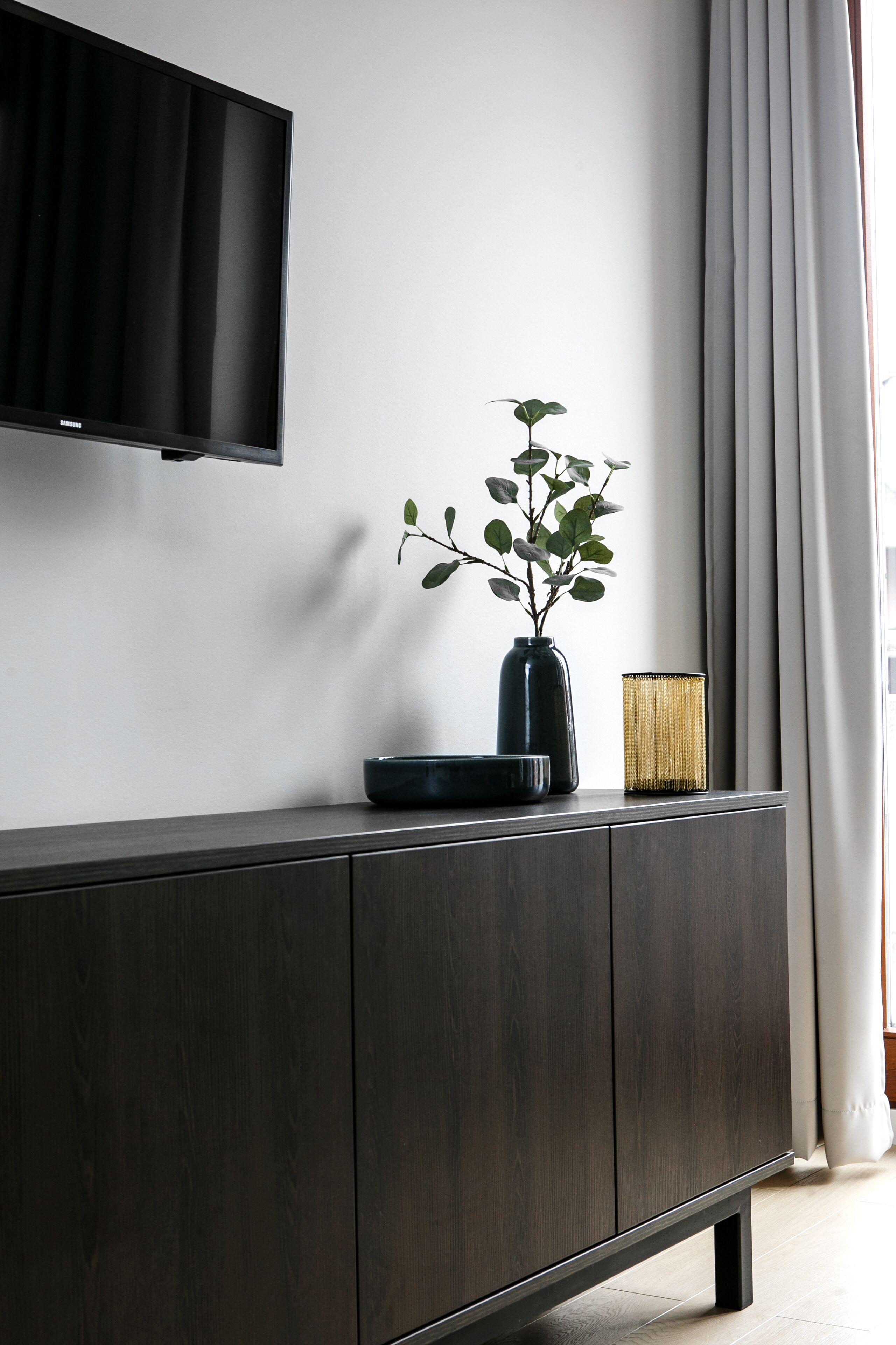 A modern living room with a dark wood cabinet, a flat-screen TV mounted on the wall, and a small plant in a vase.