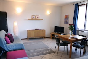 A living room with a sofa, a TV, a dining table with chairs, a cabinet, and a vase with flowers.
