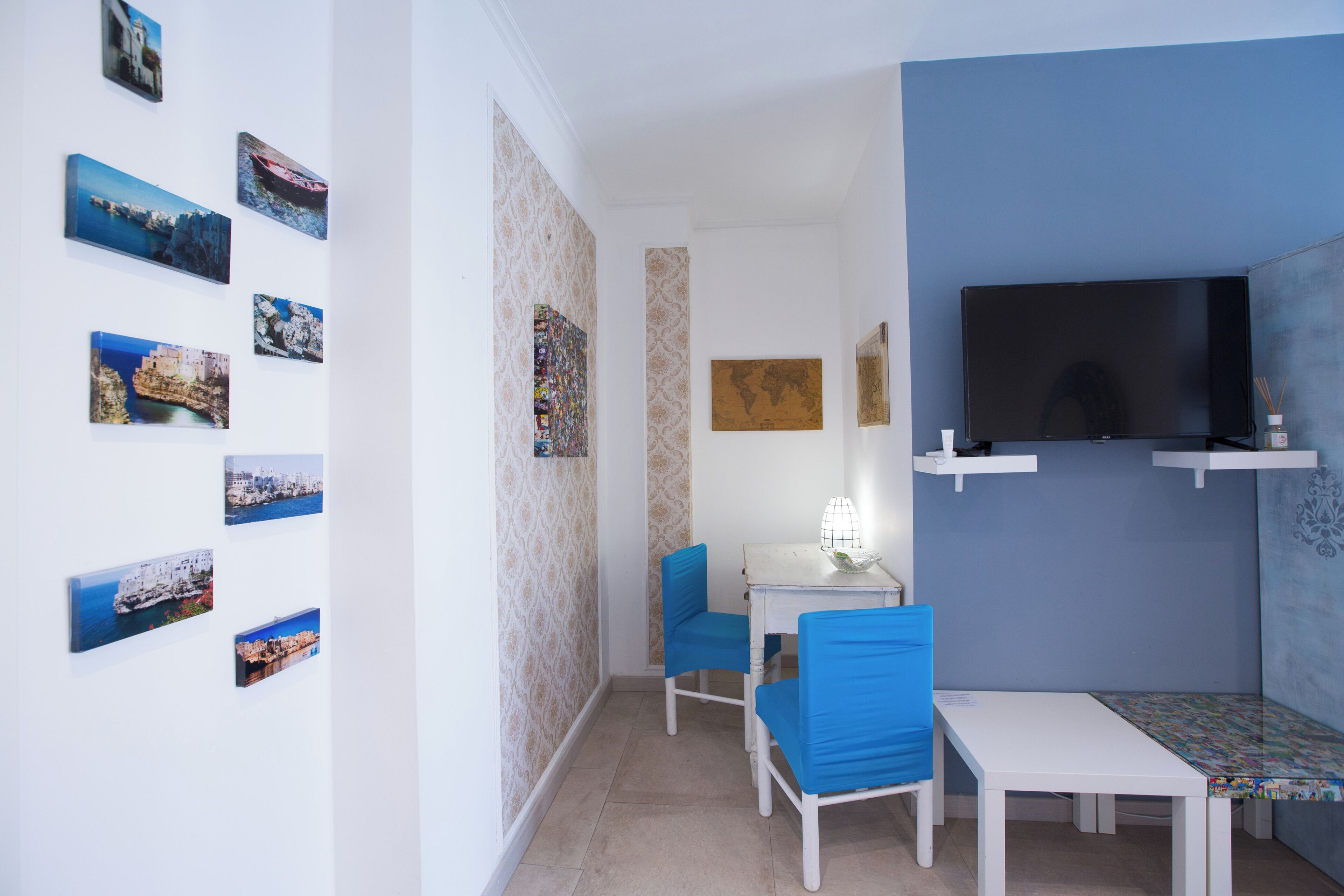 Una habitación pequeña con una mesa blanca, dos sillas azules, un televisor y varias fotos enmarcadas en la pared.