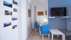 A small room with a white table, two blue chairs, a TV, and several framed pictures on the wall. - Room-versomare - Room Maestrale Extra Be (Polignano a Mare)