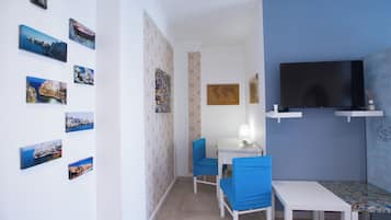 Una habitación pequeña con una mesa blanca, dos sillas azules, un televisor y varias fotos enmarcadas en la pared.
