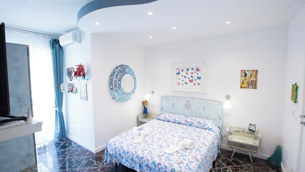 A bedroom with a bed, a mirror, a window with curtains, a bedside table, and a wall with artwork. - Room-versomare - Room Maestrale Extra Be (Polignano a Mare)