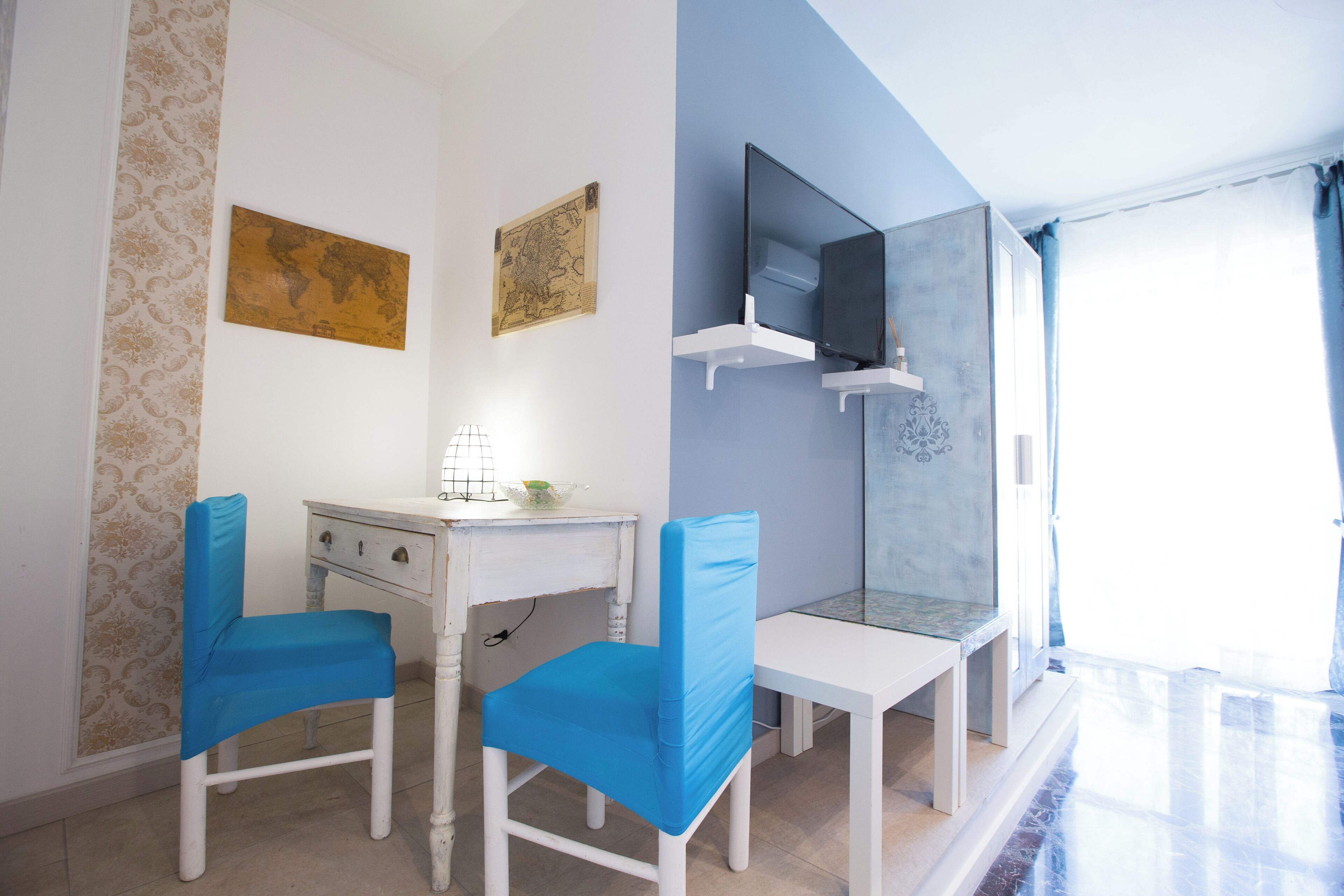 Una habitación con un escritorio blanco, dos sillas azules, un mapa mundial y una televisión sobre una estantería blanca.