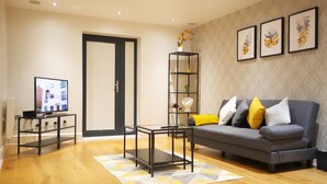 A modern living room with a grey sofa, a coffee table, a TV on a stand, a bookshelf, and a patterned rug. - The Elm Serviced Apartments (Camberley)