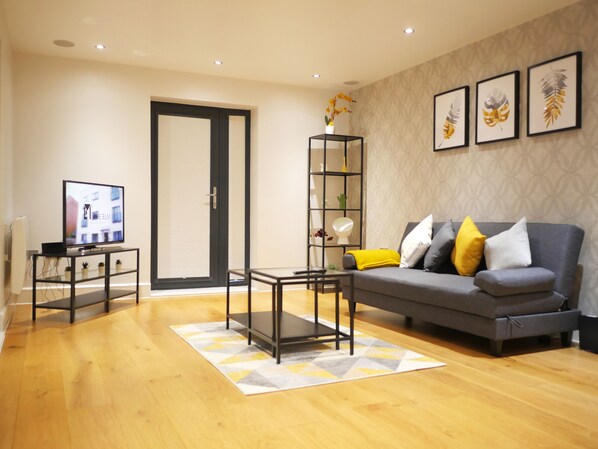 A modern living room with a grey sofa, a coffee table, a TV on a stand, a bookshelf, and a patterned rug.