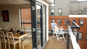 A balcony with a dining area, a sliding glass door, and a view of a brick building. - The Elm Serviced Apartments (Camberley)
