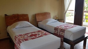 A room with two beds, each with a wooden headboard and a patterned bedspread. There is a small table with a lamp and a bottle on it. The room has a sliding glass door leading to an outdoor area. - Kampung Lumbung Boutique Hotel (Batu)