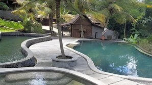 A pool area with a wooden deck, stone borders, and a small building in the background. - Kampung Lumbung Boutique Hotel (Batu)
