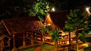 A model of a traditional wooden house with a tiled roof, surrounded by palm trees and a paved pathway. - Kampung Lumbung Boutique Hotel (Batu)