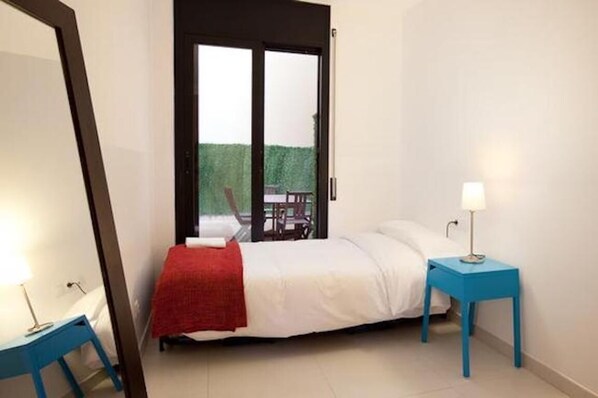 A bedroom with a bed, a red blanket, a blue nightstand with a lamp, and a sliding glass door leading to an outdoor area. - Spacious Apartment for Families Near Park Guell (Barcelona)