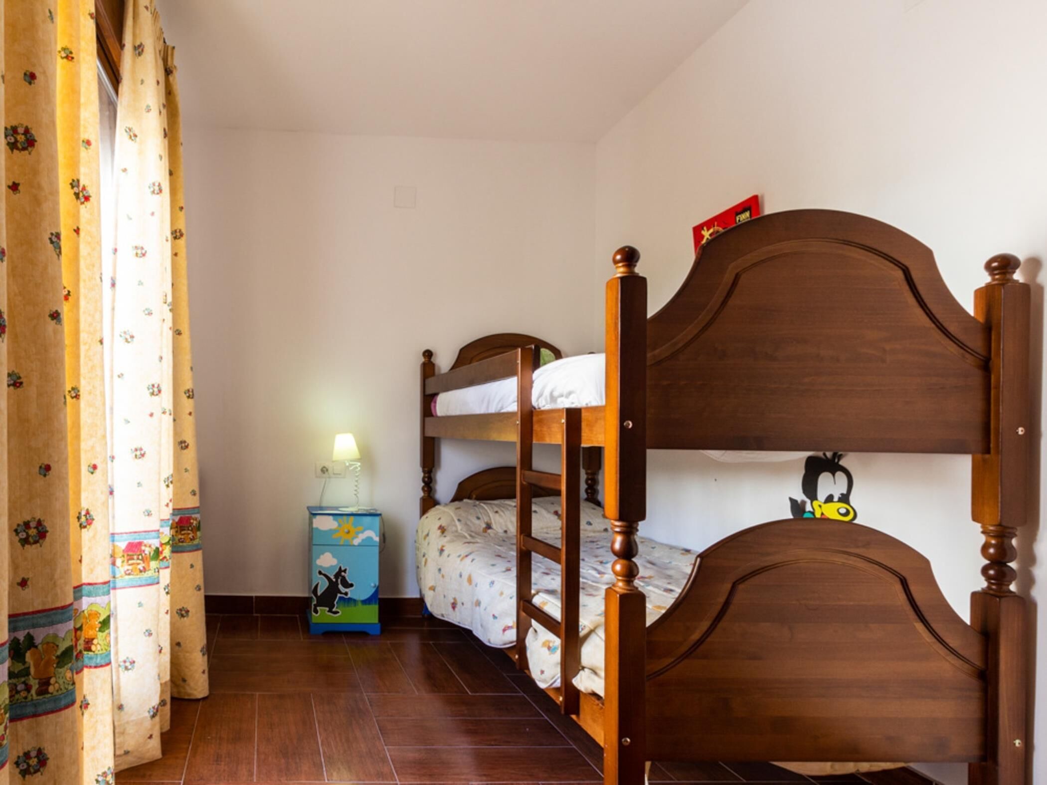 Ein Schlafzimmer mit zwei Etagenbetten, einem Nachttisch mit Lampe und einer kleinen Kommode mit Cartoon-Motiv.