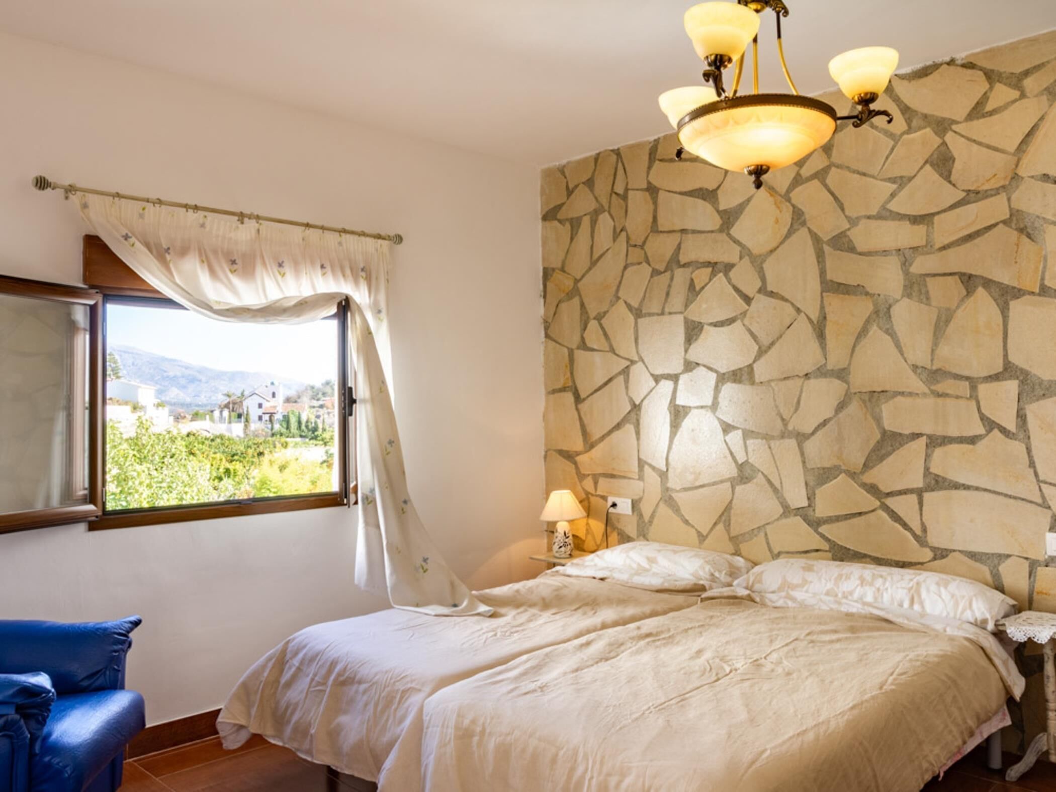 A bedroom with a stone wall, a bed, a nightstand with a lamp, a window with curtains, and a view of greenery outside.