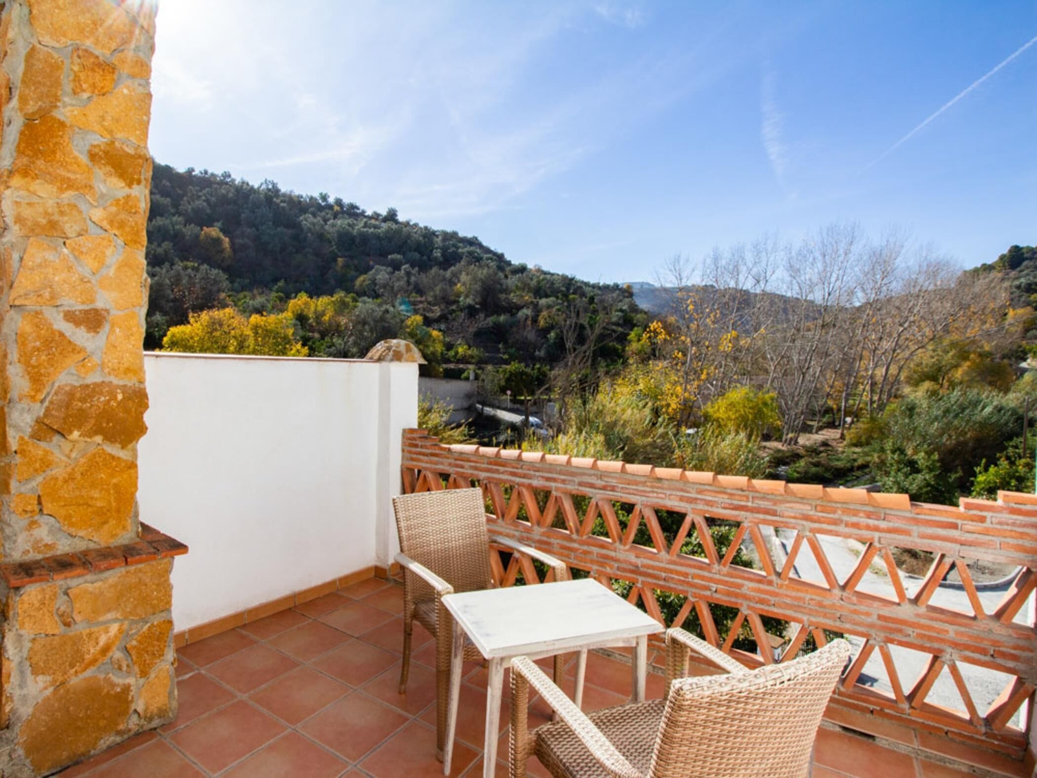 A balcony with wicker furniture, a stone wall, and a view of a hilly landscape.