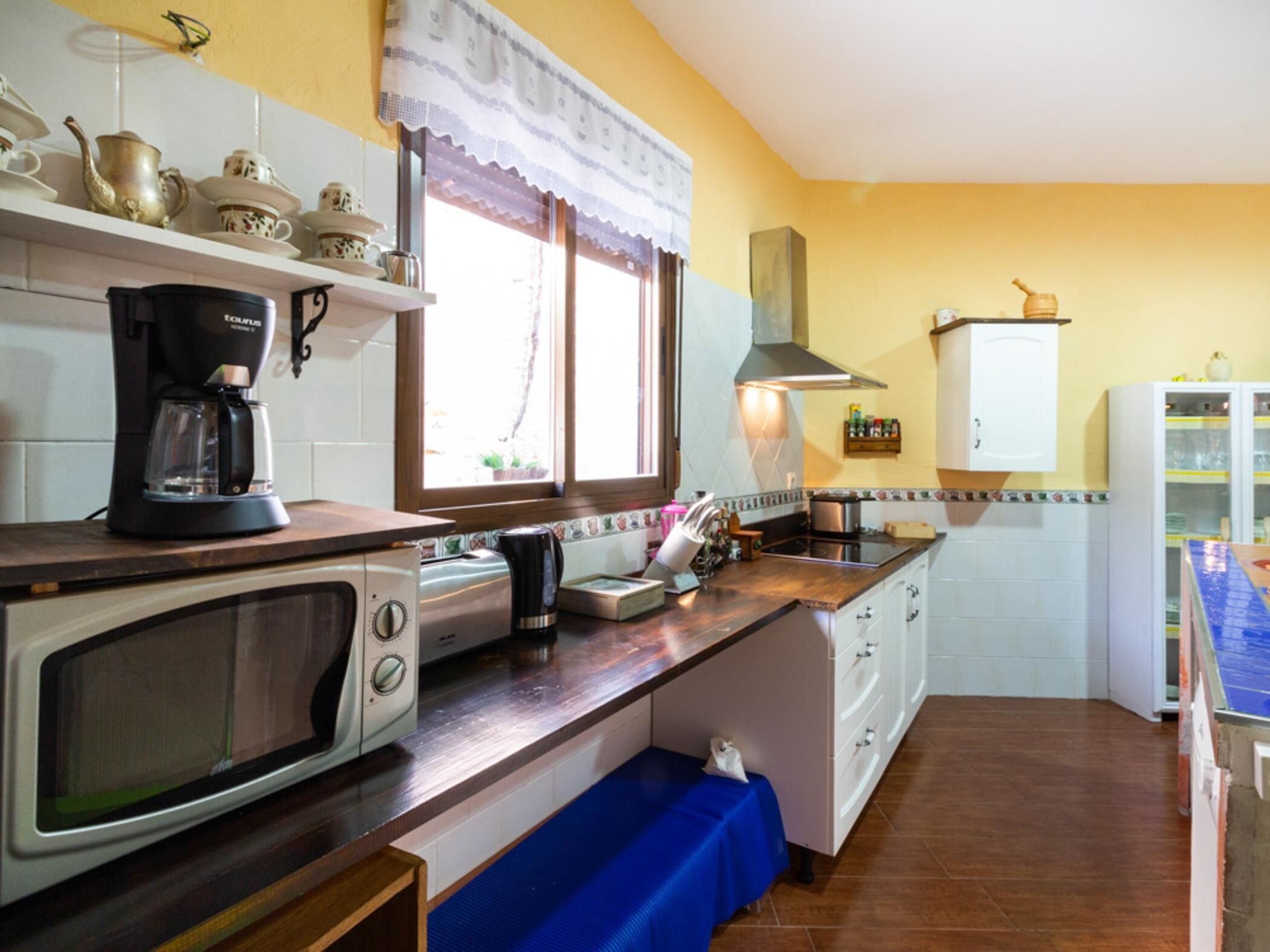 A compact kitchen with a microwave, coffee maker, and a stove. There is a window with a curtain, a sink, and a cabinet with dishes.