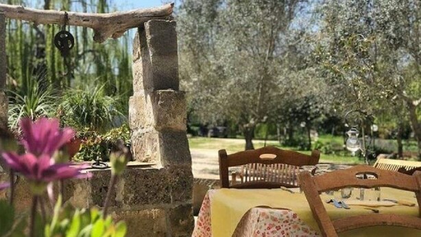 A rustic outdoor seating area with stone walls, a wooden table with a floral tablecloth, and a chair. - APARTMENT 2 ON GARDEN (UGGIANO LA CHIESA)