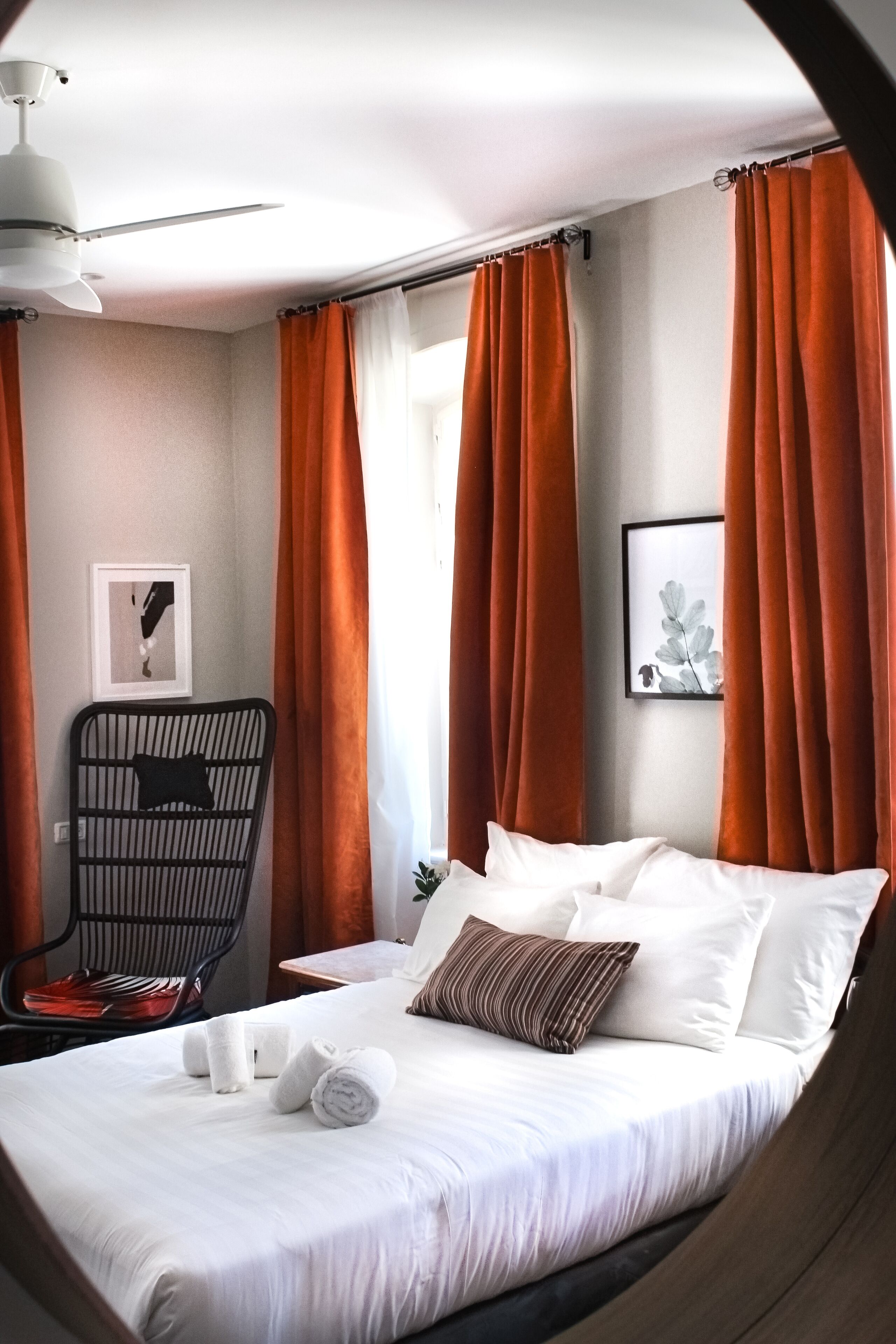A neatly made bed with white linens, a black chair with a cushion, and orange curtains.