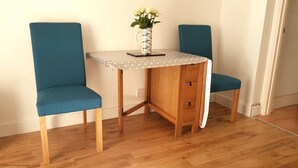 Een klein houten bureau met een witte tafelkleed met stippen en een vaas witte bloemen. Er staan twee blauwgroene stoelen met houten poten.