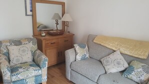 Een woonkamer met een houten dressoir, twee gestreept fauteuils, een grijze bank met bloemkussens, en een spiegel boven het dressoir.