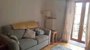 A living room with a grey sofa, a small wooden side table, a lamp, and a sliding glass door leading to an outdoor area.