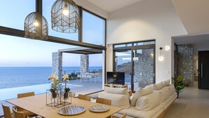 Amplia sala de estar con una gran mesa, asientos cómodos y un televisor. Se aprecia una pared de piedra y vistas al mar a través de amplias ventanas.