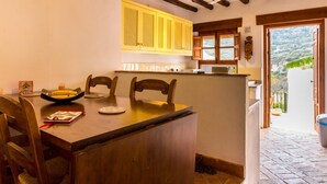 A dining area with a wooden table and chairs, a kitchen with yellow cabinets, and a view of the outdoors through a door. - Cosy restored rural house (Albuñuelas)