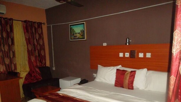 A hotel room with a large bed, a nightstand, a desk, and a painting on the wall. - City Crown Hotels Limited (Port Harcourt)