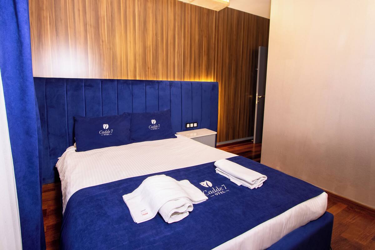 Camera d'albergo con un letto grande, testiera blu e copripiumino blu. Ci sono due cuscini con il logo dell'hotel, un asciugamano piegato e un comodino con una lampada.
