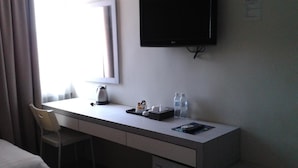 Habitación de hotel con escritorio, una silla, televisor de pantalla plana, ventana con cortinas y mesita de noche con control remoto.