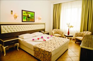 A hotel room with a large bed, a bedside table, a chair, a small table with a vase of flowers, and a television on the wall. - Phoenix Sun Hotel (Bodrum)