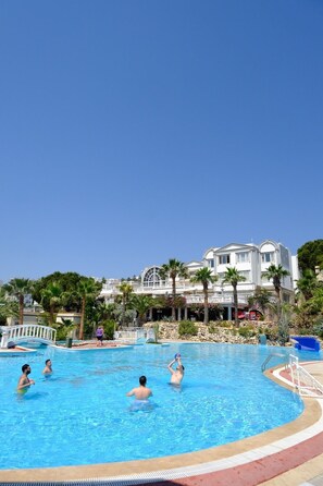 A swimming pool with people in it, surrounded by palm trees and a large building in the background. - Phoenix Sun Hotel (Bodrum)