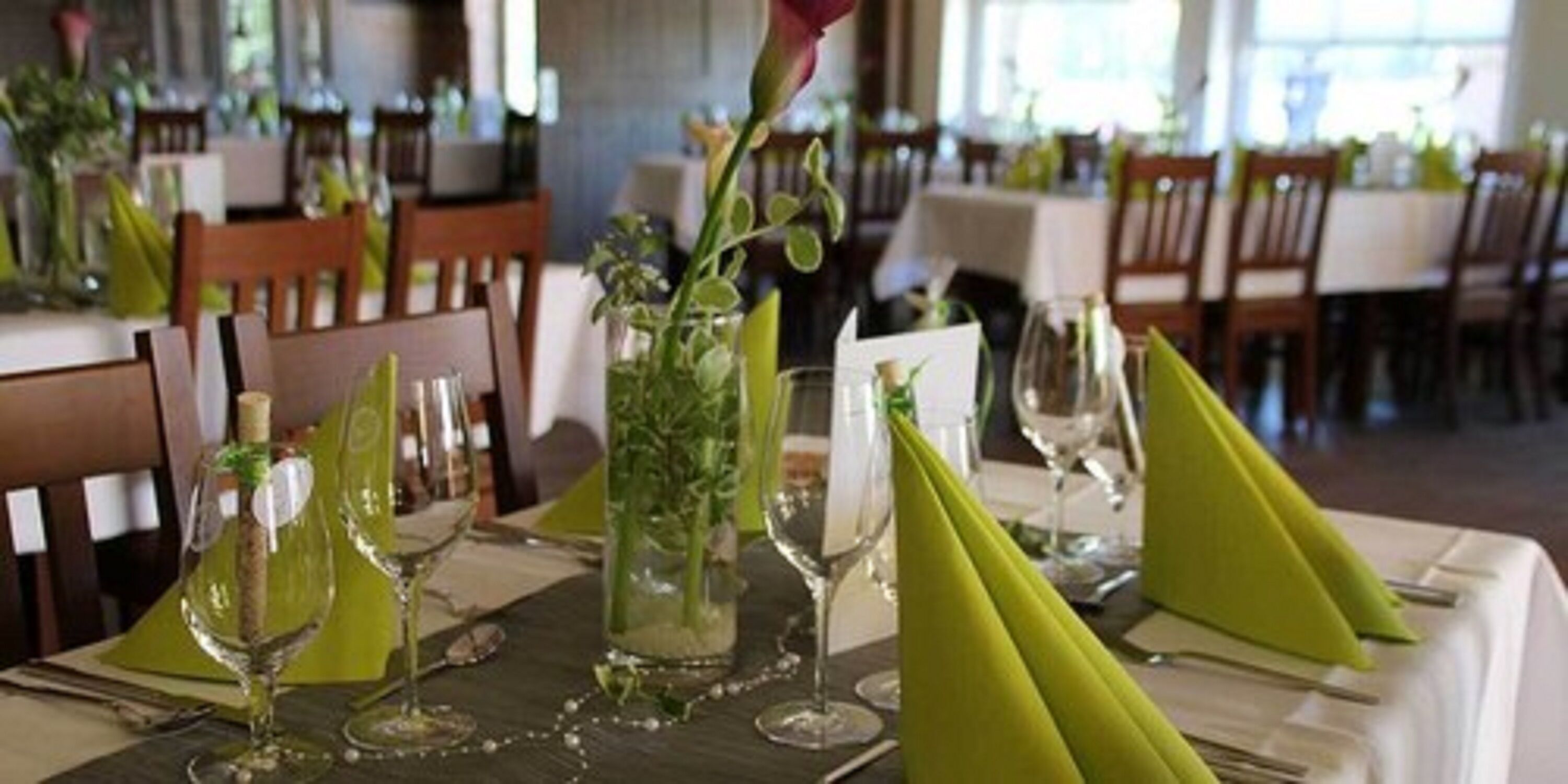 Una mesa de comedor con copas de vino, un florero con follaje y servilletas verdes.