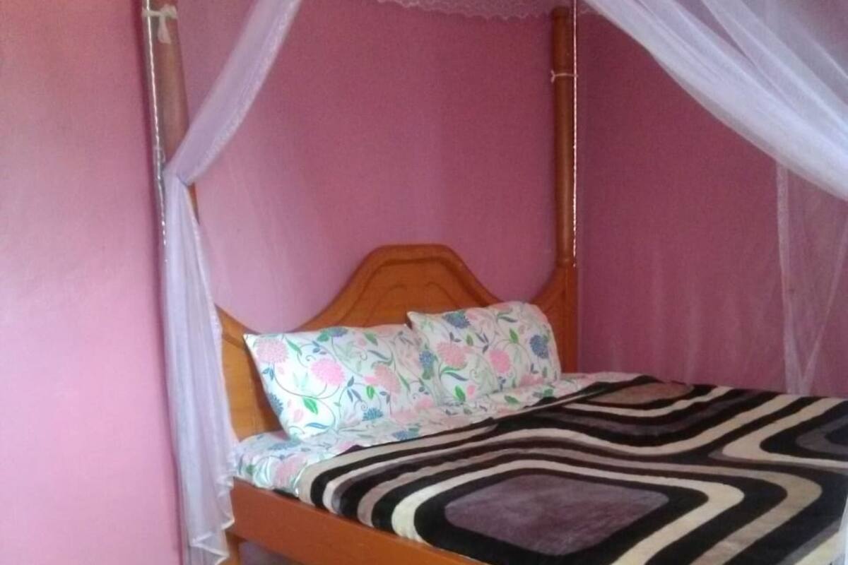 A single bed with a wooden frame and a canopy, a patterned bedspread, and a pair of slippers on the floor.