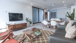 Un salon moderne doté d’un téléviseur à écran plat, d’un canapé, d’une table basse avec des magazines, d’un coin repas avec des chaises et d’une grande fenêtre.