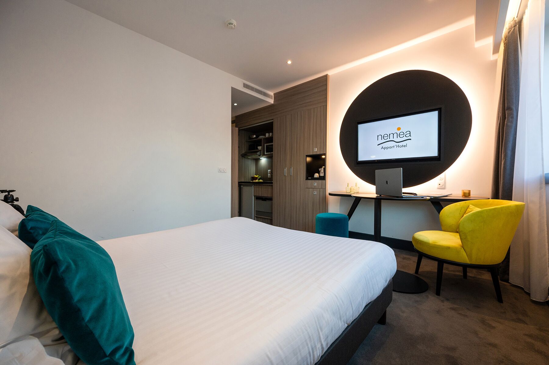 A modern hotel room with a bed, a desk with a laptop, a TV, and a yellow chair.