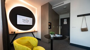 A reception area with a modern design, featuring a large circular light fixture, a yellow armchair, a small table with a plant, and a flat-screen TV displaying the hotel name.