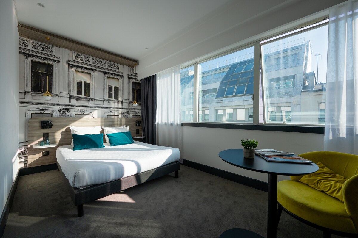 Habitación de hotel moderna con una cama grande, una mesita redonda pequeña, un sillón amarillo y vistas de la ciudad a través de ventanas amplias.