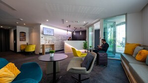 A modern hotel lobby with a reception desk, seating area, and a large window offering a view of the street.