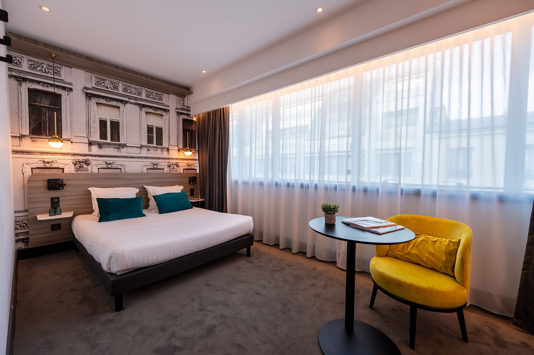 A hotel room with a bed, a small table, a yellow armchair, and a large window with sheer curtains.