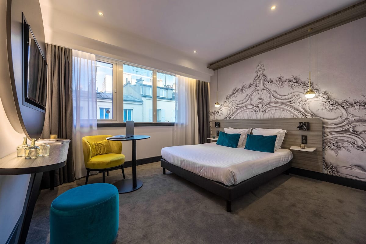 A hotel room with a bed, a desk, a chair, and a view of the city through large windows.