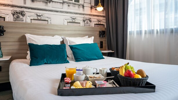 A hotel room with a bed, two pillows, a tray with breakfast items, a fruit basket, and a view of a building with classical architecture.