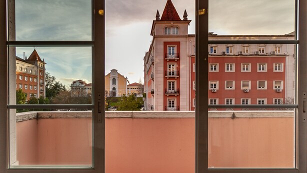 A view through a window of a building with a pink wall and a red-roofed tower. - Residencial Horizonte (Lisbon)